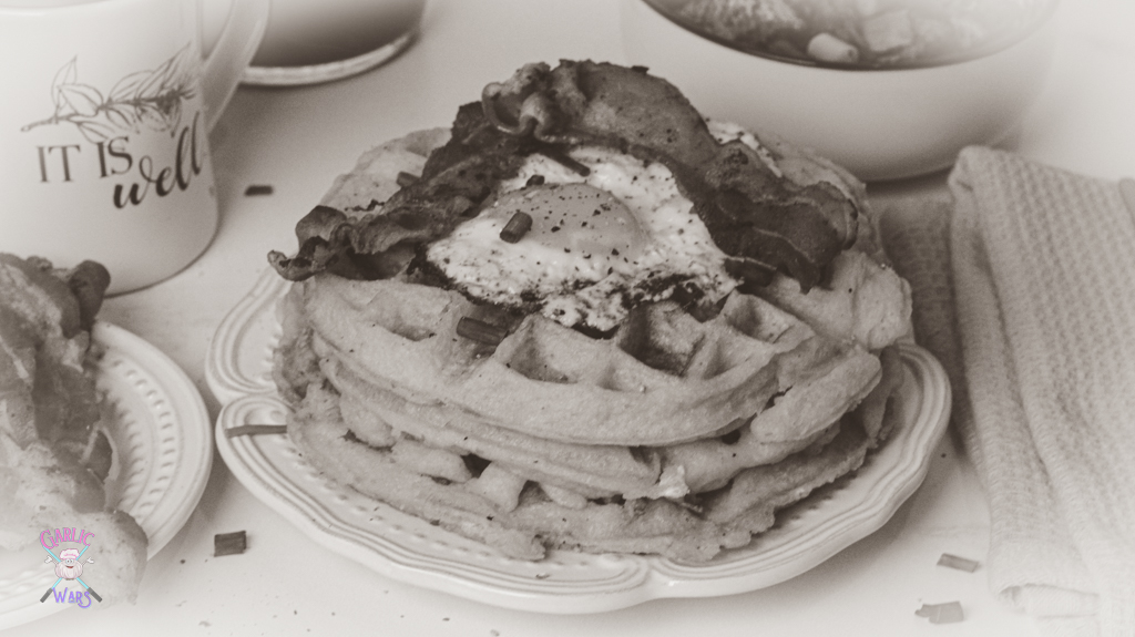 A plate with waffles, topped with bacon and a fried egg, with a plate of bacon on the side, and a coffee mug that reads "it is well." A black and white picture to represent the style of WandaVision.
