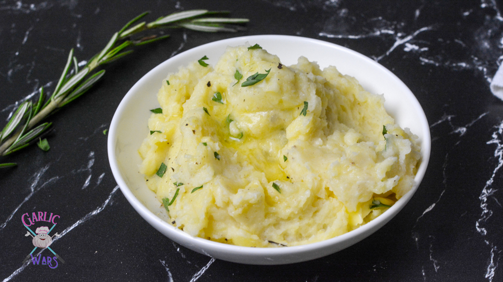 A small white bowl of mashed potatoes with melted butter on a black marble countertop, and a sprig of rosemary.
