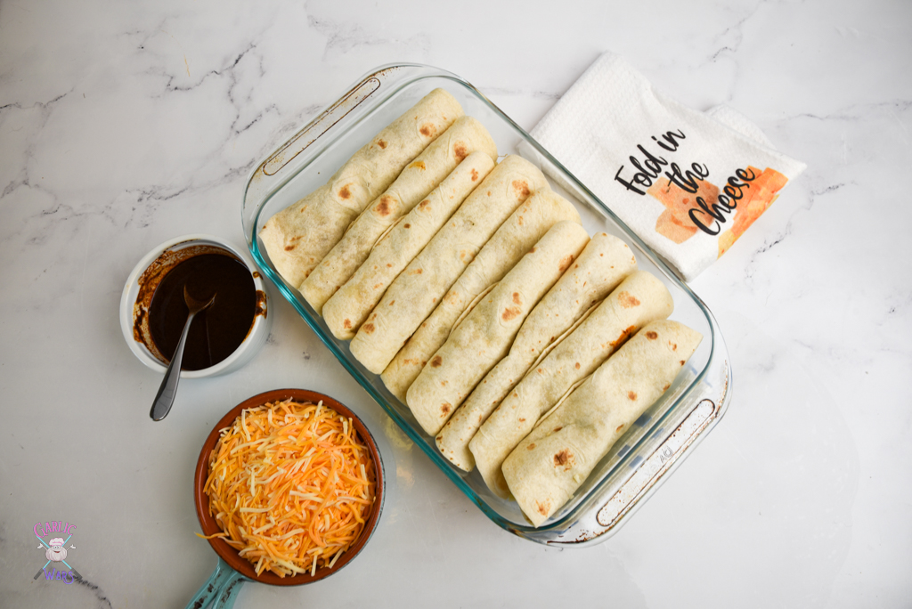 large baking sheet with tortillas, enchilada sauce on the side, cheese on the side, and a tea towel that reads "fold in the cheese"