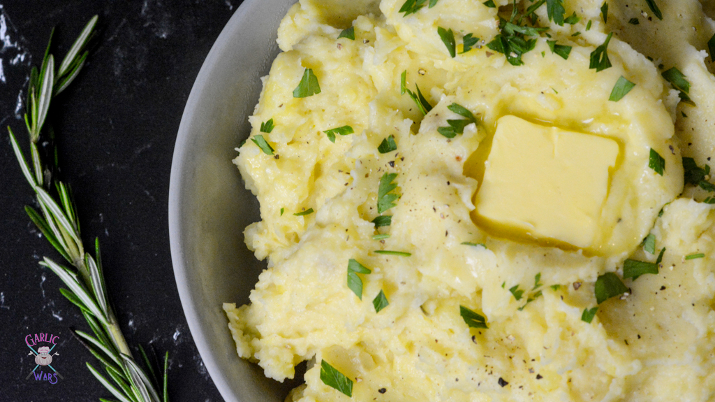 A close up bowl of mashed potatoes, topped with melting butter, fresh parsley, and cracked black pepper.