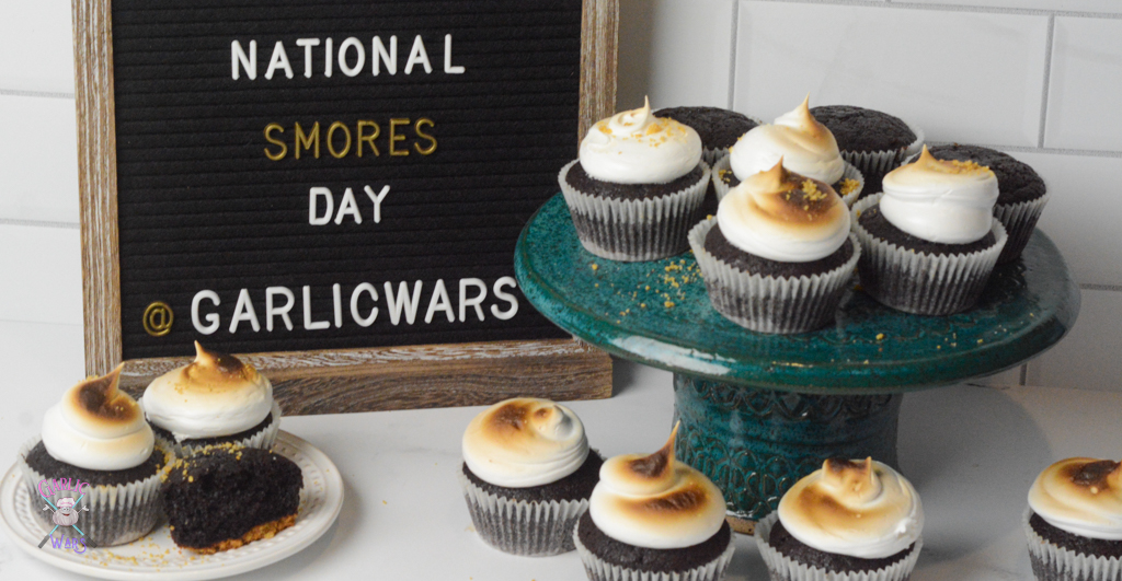 cupcakes on a teal cake stand, with more on the surrounding table, with a black letter board sign that reads "national smores day"