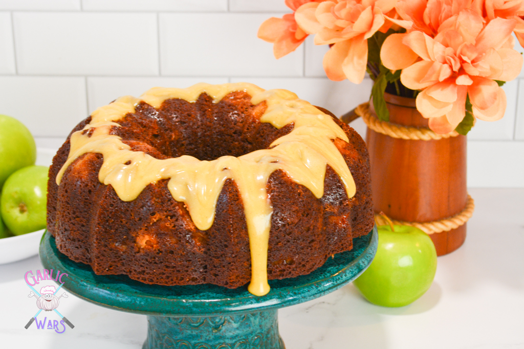 dark brown bundt cake on a teal cake stand with a golden blonde colored glaze dripping down. Background has green apples and orange flowers.