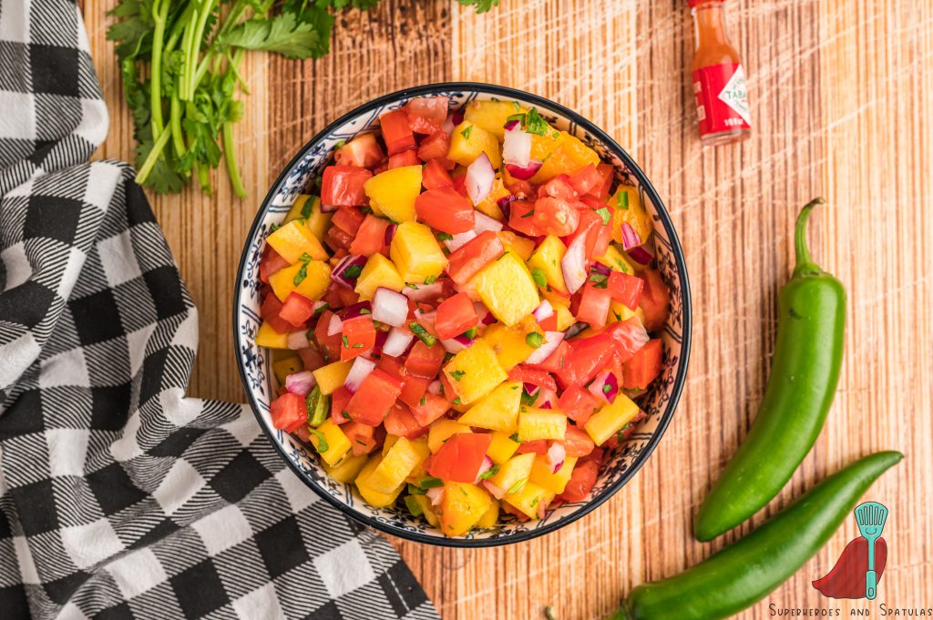 peach salsa in a small blue and white bowl with a black and white towel, jalapenos, cilantro, and tabasco